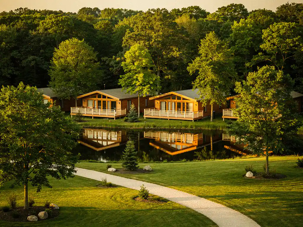 Luxe glamping resort met houten lodges en overdekte veranda's weerspiegeld in een rustige vijver, omgeven door weelderig groen bij gouden uurtje.