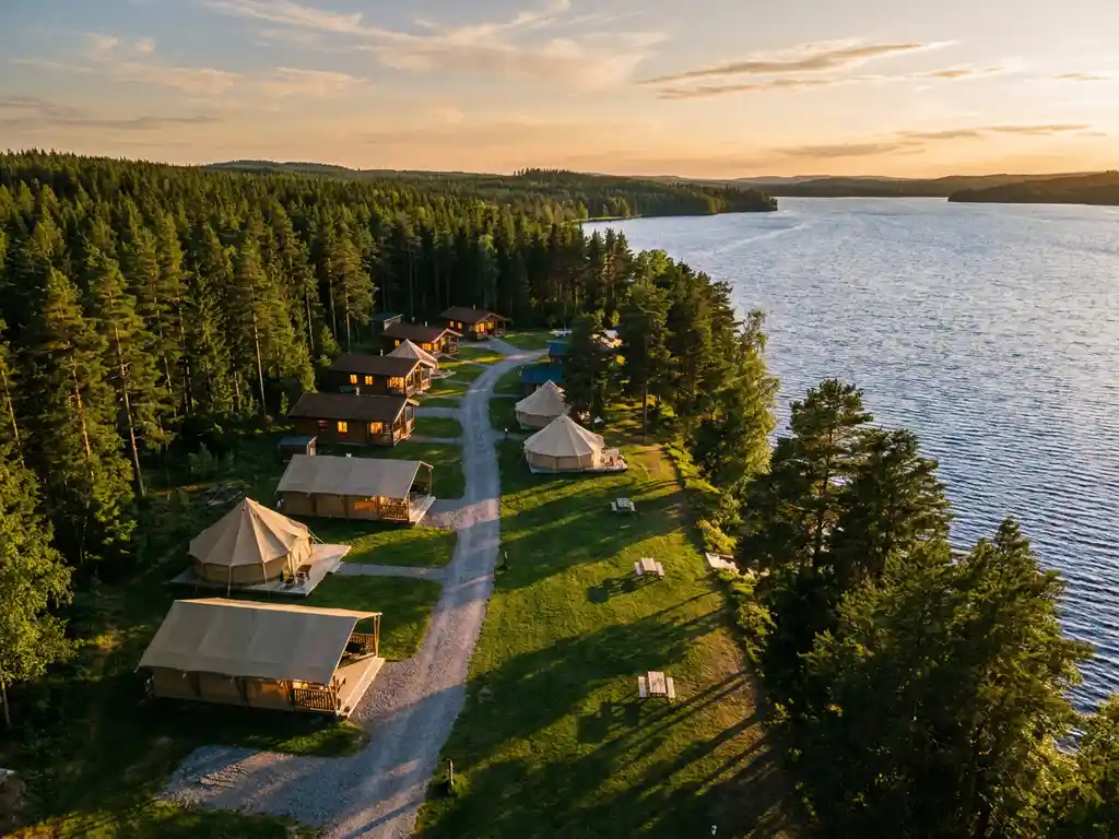 Luchtfoto van een luxe glamping met tenten en houten lodges langs een grindpad, omgeven door dennenbos en een meer in gouden middaglicht.