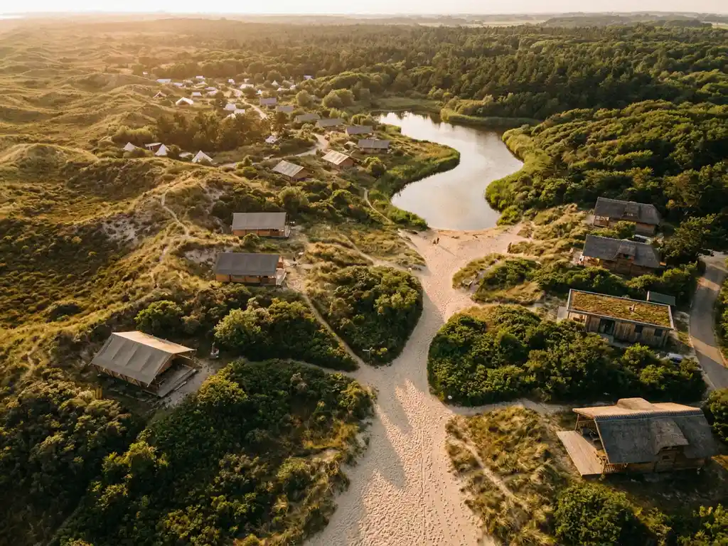 Luchtfoto van een Nederlands vakantiepark met glamping lodges en chalets tussen groene duinen en kustbos, kronkelend zandpad richting water.