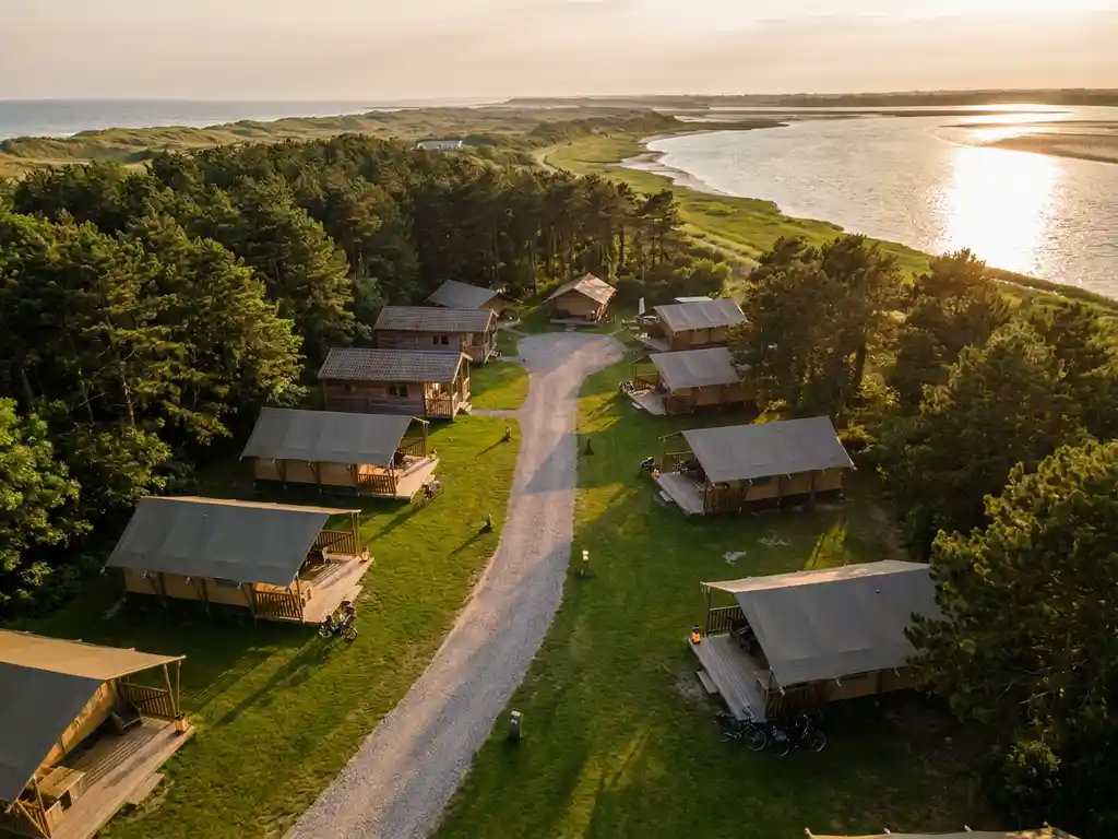 Luchtfoto van glamping-tenten en houten lodges langs kronkelend grindpad, omgeven door dennenbossen aan de Nederlandse kust bij gouden uur.