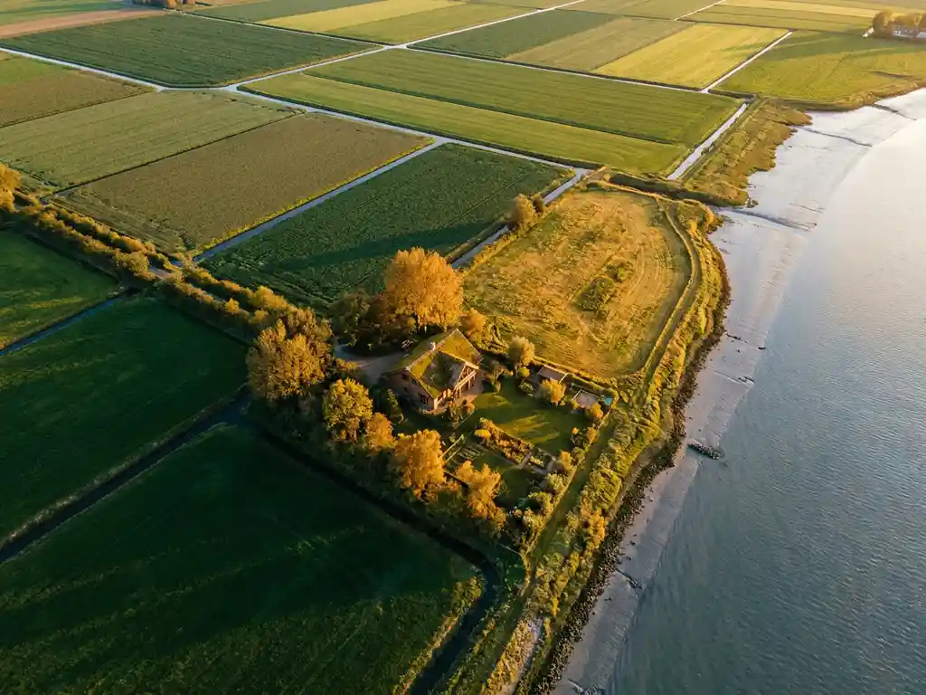 Luchtfoto van Zeeuws polderlandschap bij gouden uur, met glooiende percelen in groen en amber en een luxe chalet tussen bomen.
